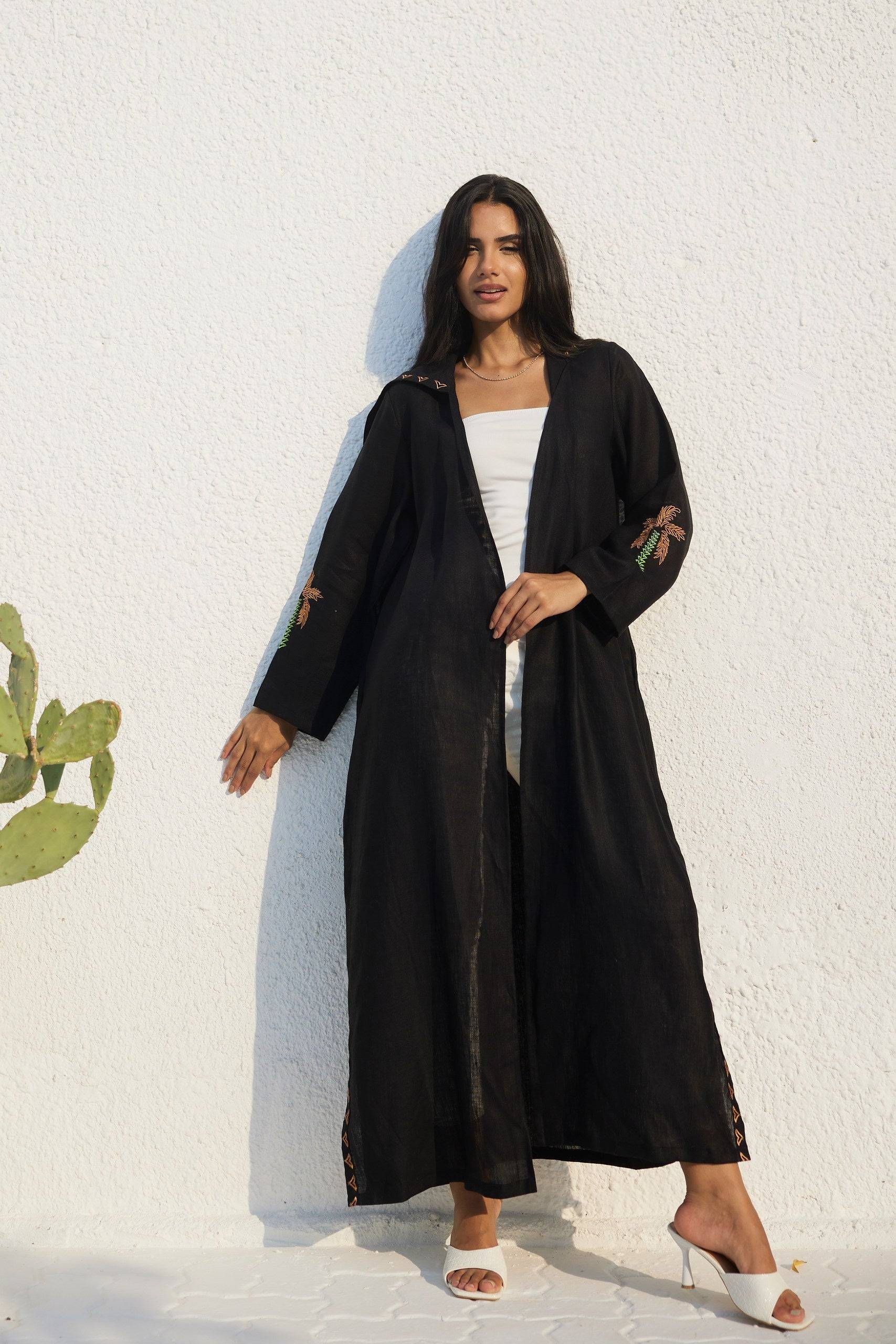 Model leaning against wall in Nakhla Hoodie Abaya with hidden pockets and palm embroidery. Made from soft organic linen.