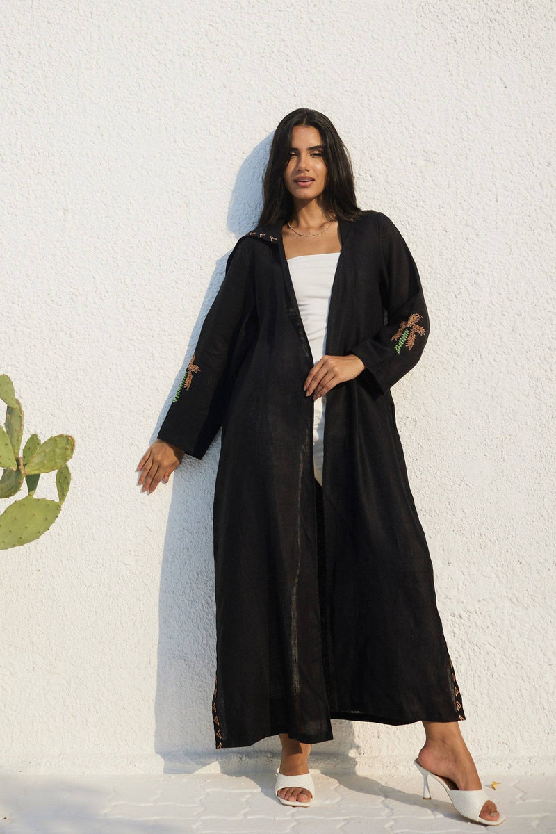 Model leaning against wall in Nakhla Hoodie Abaya with hidden pockets and palm embroidery. Made from soft organic linen.