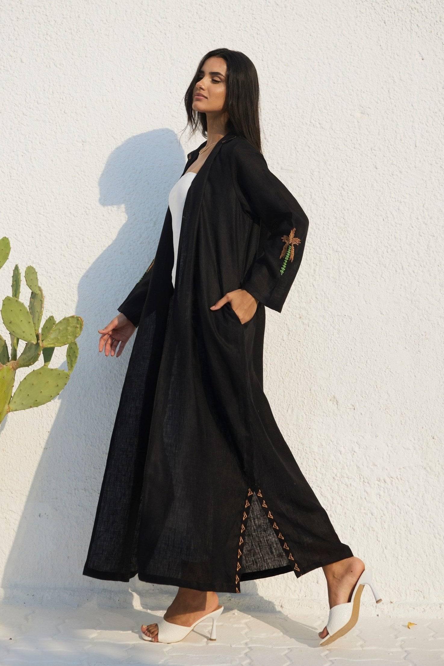 Flowy Nakhla Hoodie Abaya in black organic linen with palm embroidery and side slits. Styled with white heels for a chic look.