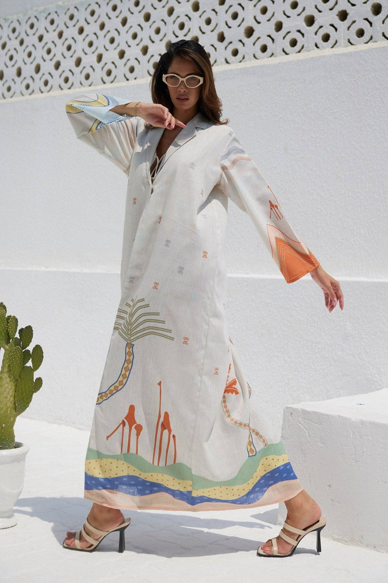 The Jeddah Horizon - Abaya & Scarf Set in white linen with colorful abstract desert and palm tree prints, styled with heels outdoors.