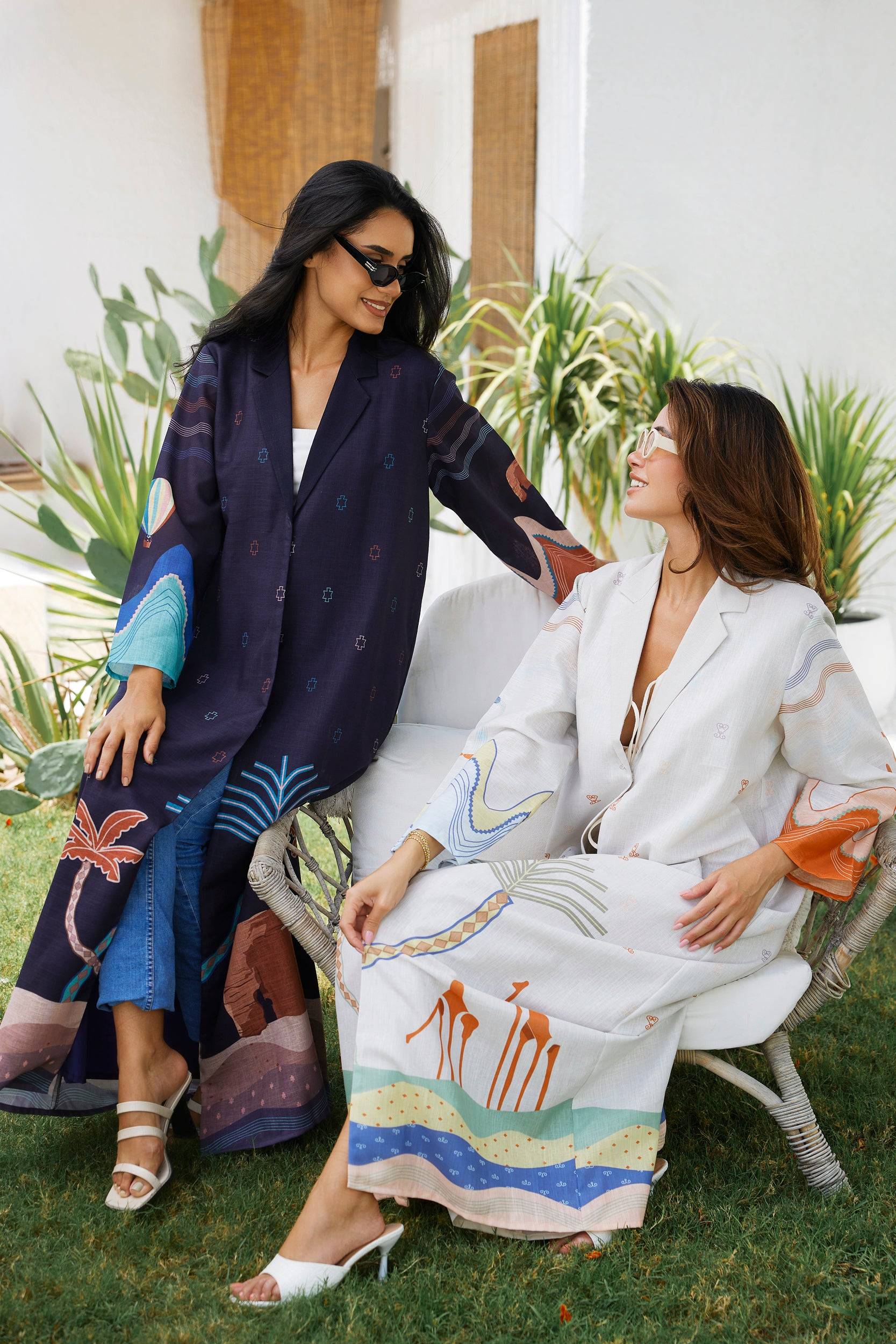 Two women wearing The Alula Horizon Blazer Abaya in navy and white versions, seated outdoors in a relaxed, stylish setting.