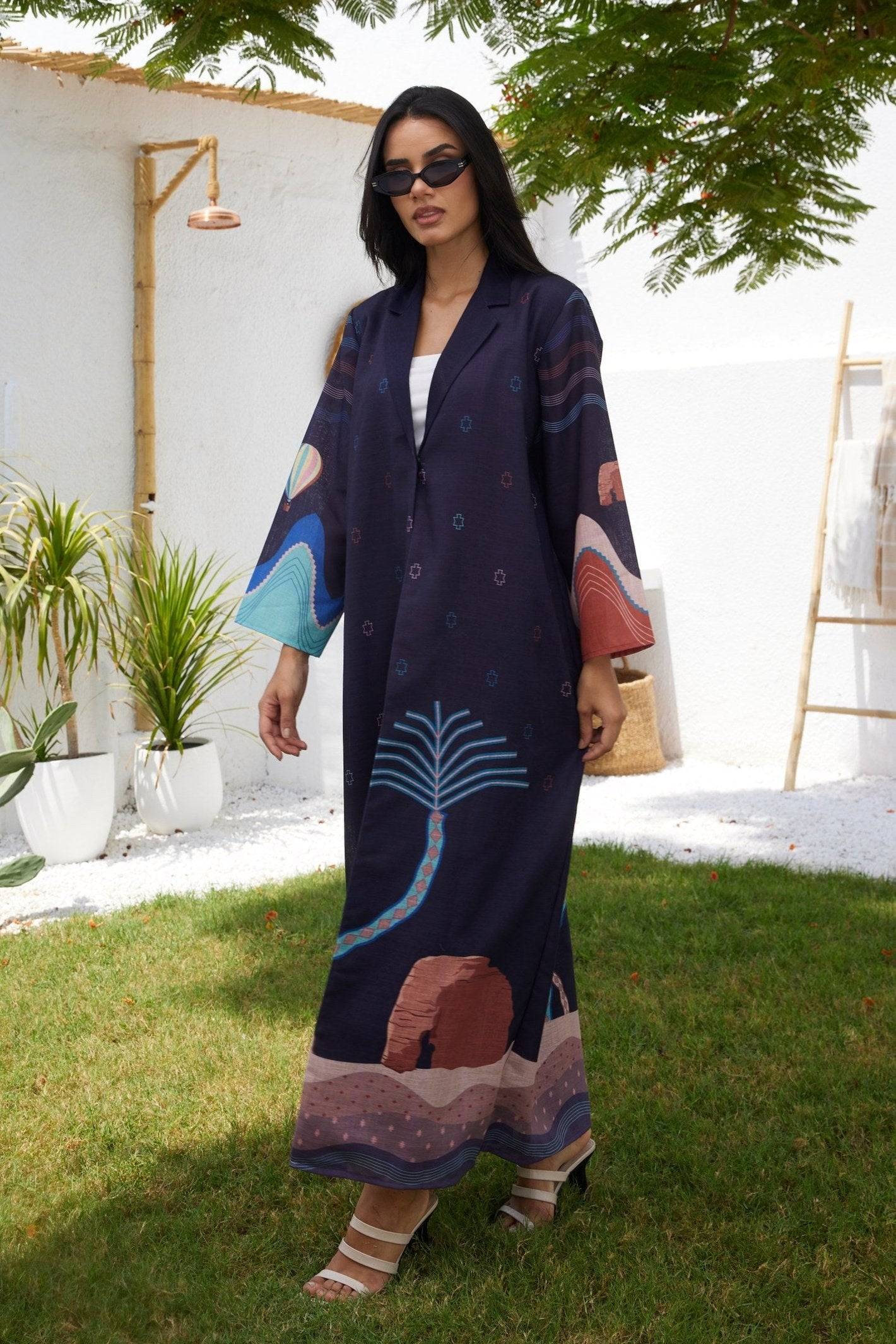 The Alula Horizon Blazer Abaya styled with white heels and sunglasses, featuring artistic desert landscape prints on a navy base.
