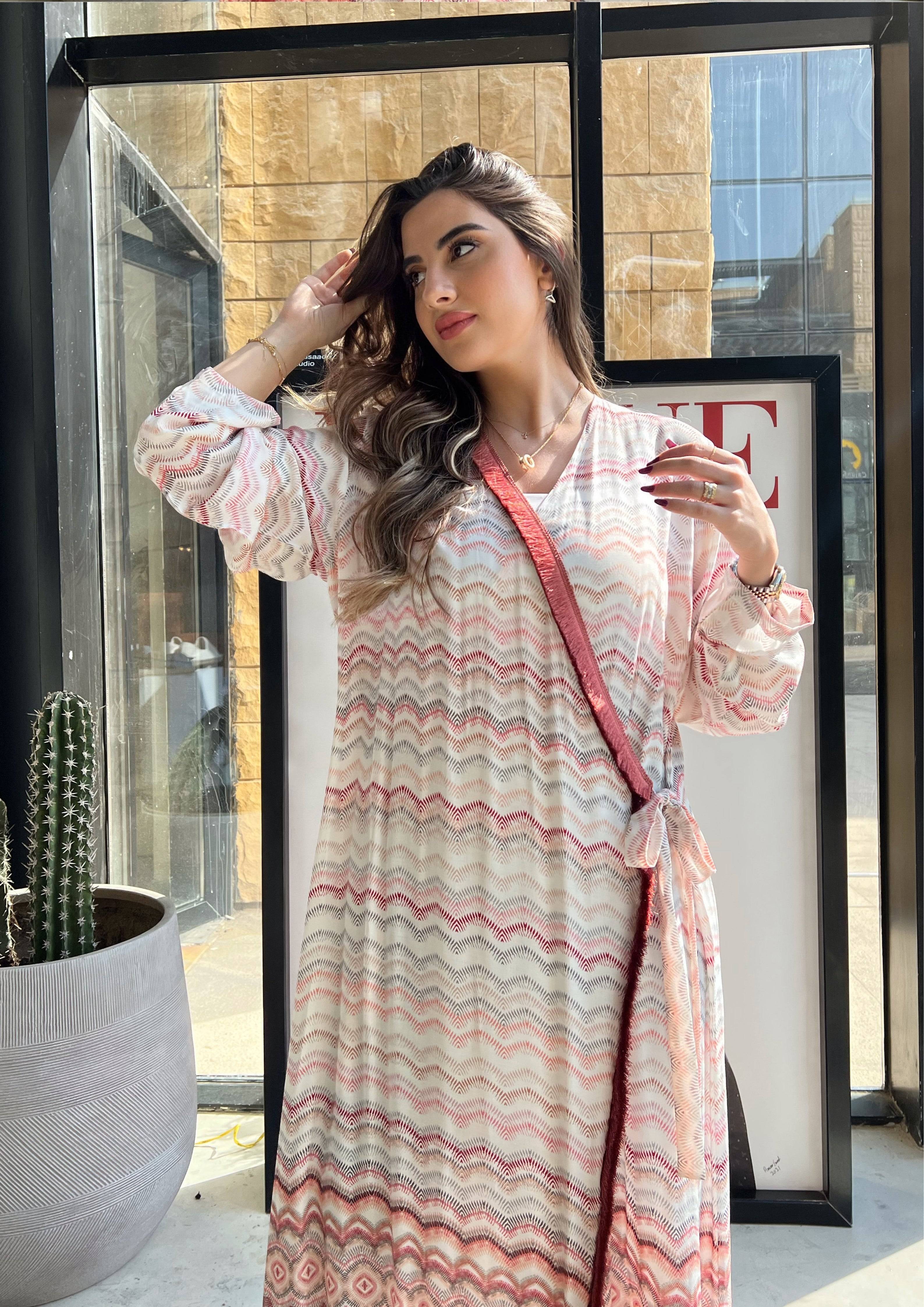 Model wearing pink print soft abaya with red velvet trim, standing near window. Saudi-made wrap kimono for chic modest outfits.