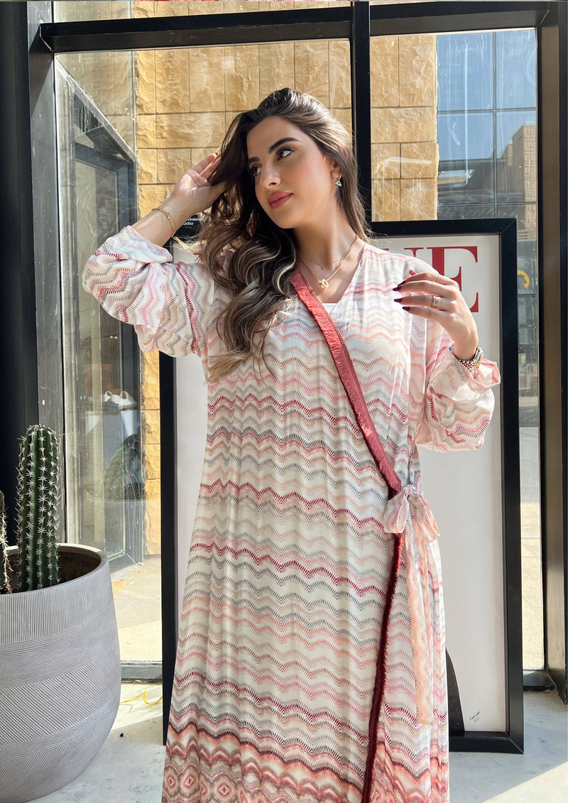 Model wearing pink print soft abaya with red velvet trim, standing near window. Saudi-made wrap kimono for chic modest outfits.