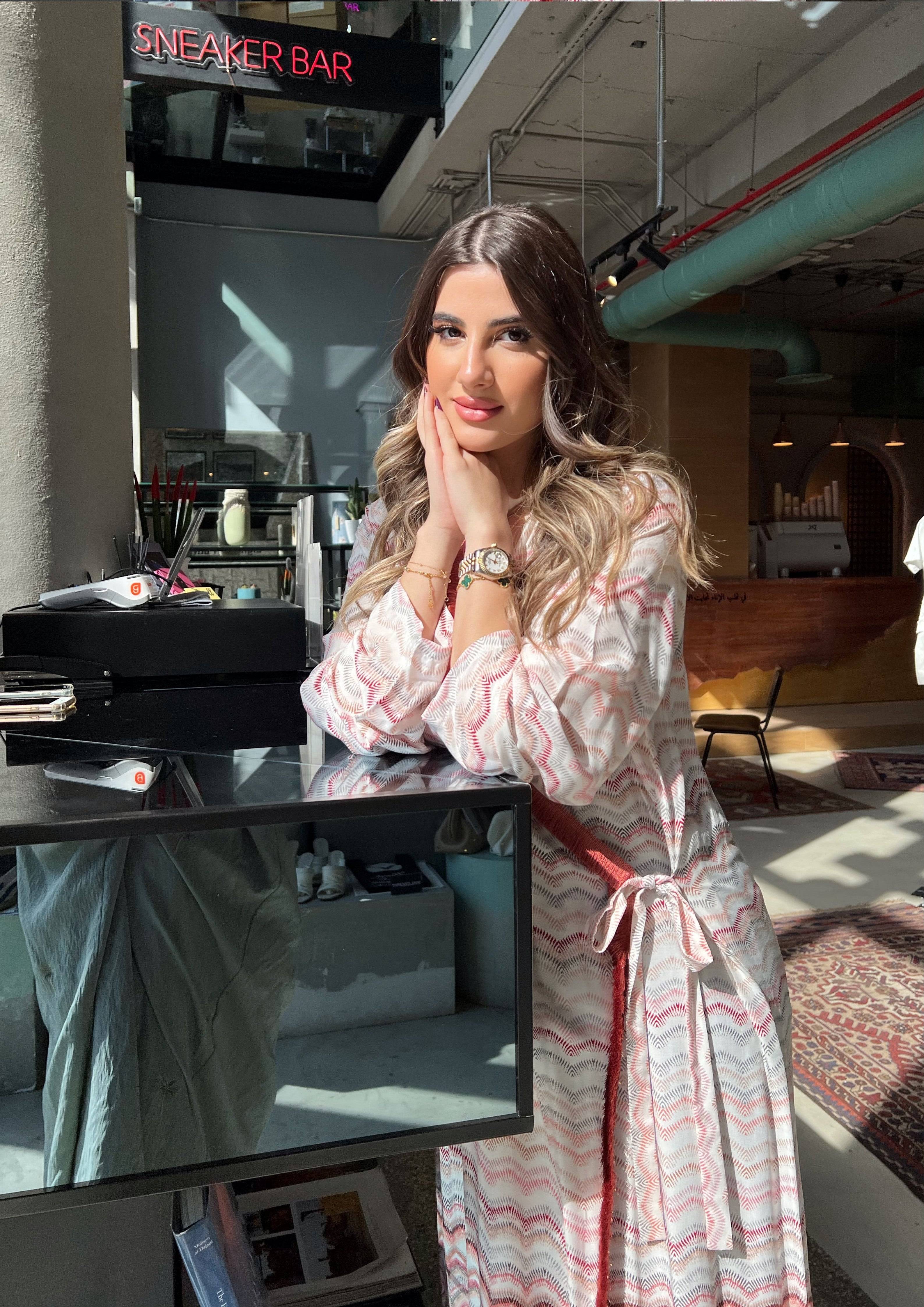 Model in pink print abaya with red velvet wrap, seated in modern cafe. Saudi-made kimono ideal for modest fashion lovers.