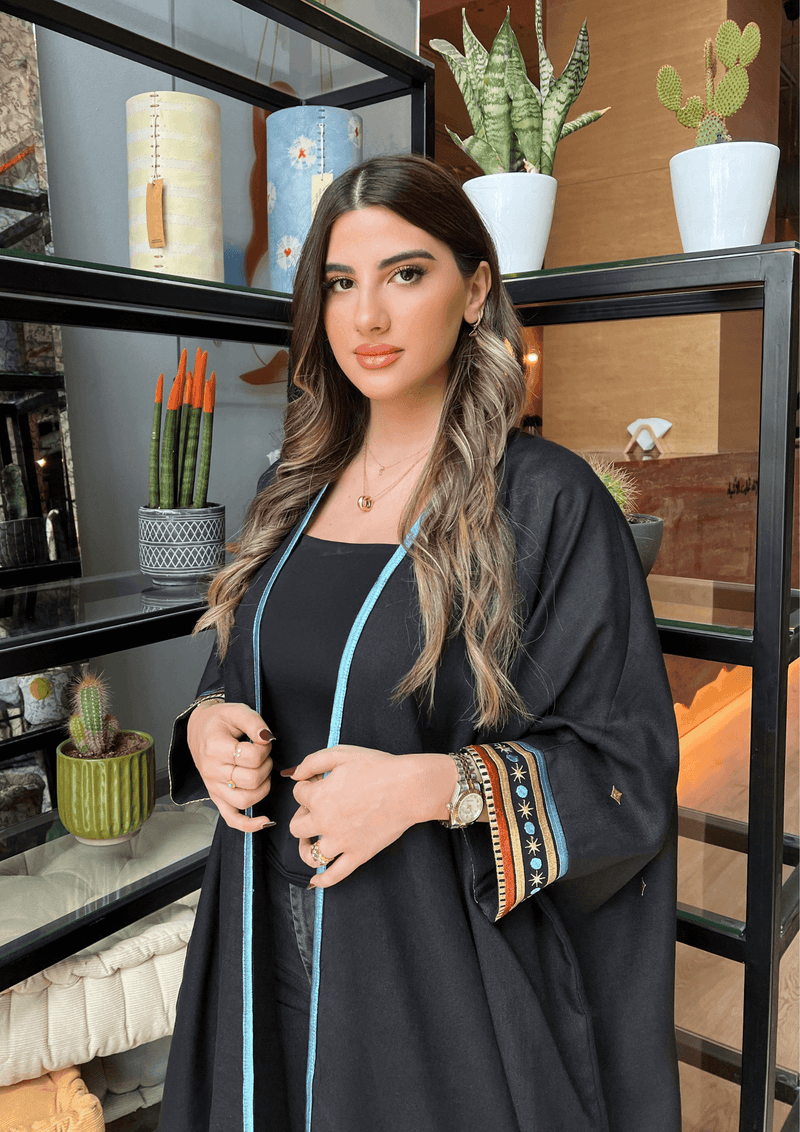 Model in Distant Galaxies - Show Stopper Back Abaya posing indoors with embroidered sleeves and blue trim, styled for modern modest fashion.
