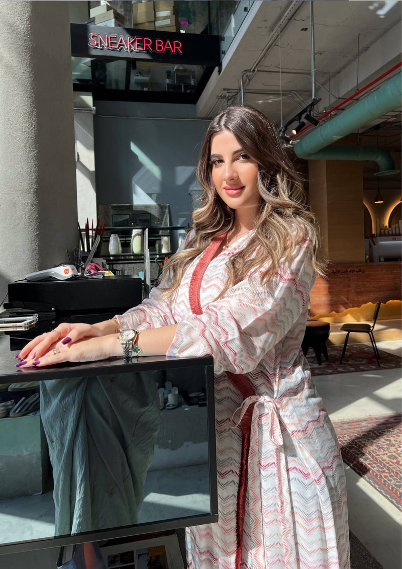 Model in pink print wrap abaya with red velvet trim, leaning on counter. Saudi-made kimono for modern modest fashion.