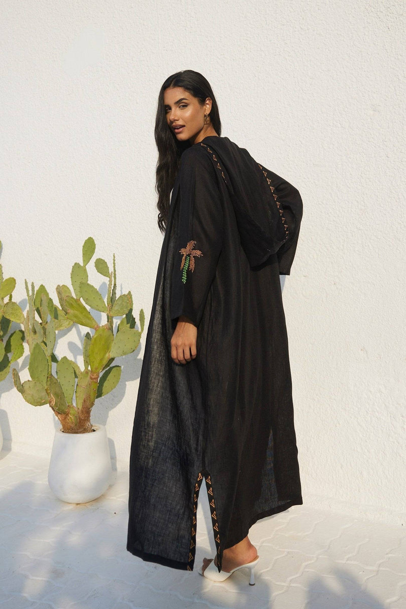 Model wearing Nakhla Hoodie Abaya in black organic linen, showcasing palm embroidery and flowing silhouette.