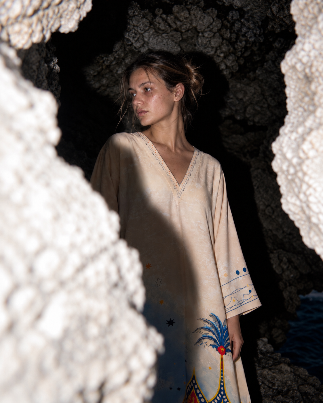 A woman wearing a cream-colored kaftan dress with colorful painted details stands inside a rocky cave, framed by white rock formations in the foreground.