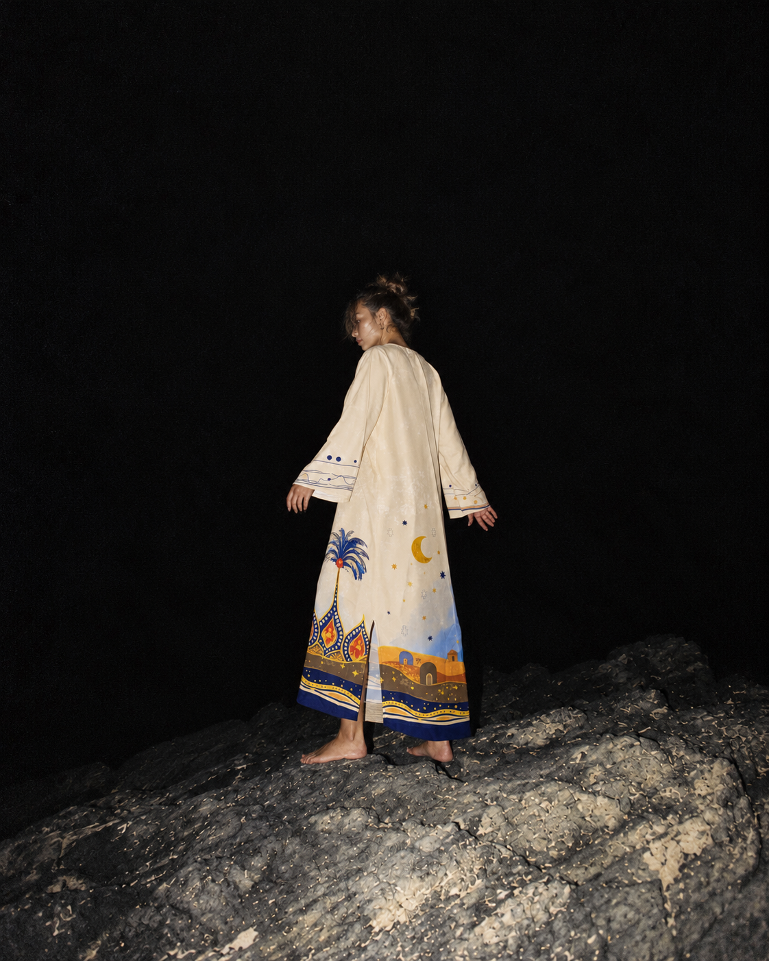A woman stands barefoot on a dark rocky surface at night, photographed from behind, wearing a long cream-colored robe with colorful desert and palm tree illustrations near the hem.