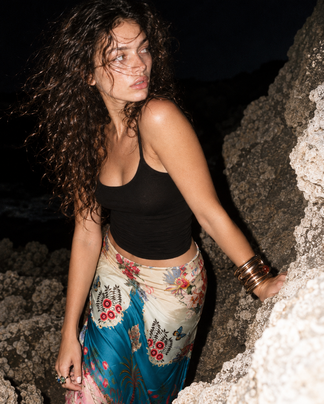 A woman posing outdoors at night against a rocky cliff, wearing a black tank top and a colorful floral wrap skirt with blue, pink, and beige tones.