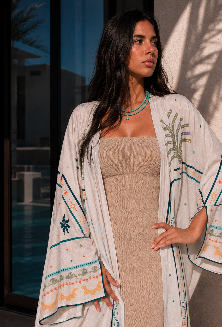 A woman wearing a beige ribbed dress and a white bohemian-style open-front kimono cover-up with teal and orange geometric patterns, standing in front of a wall with palm leaf shadows.