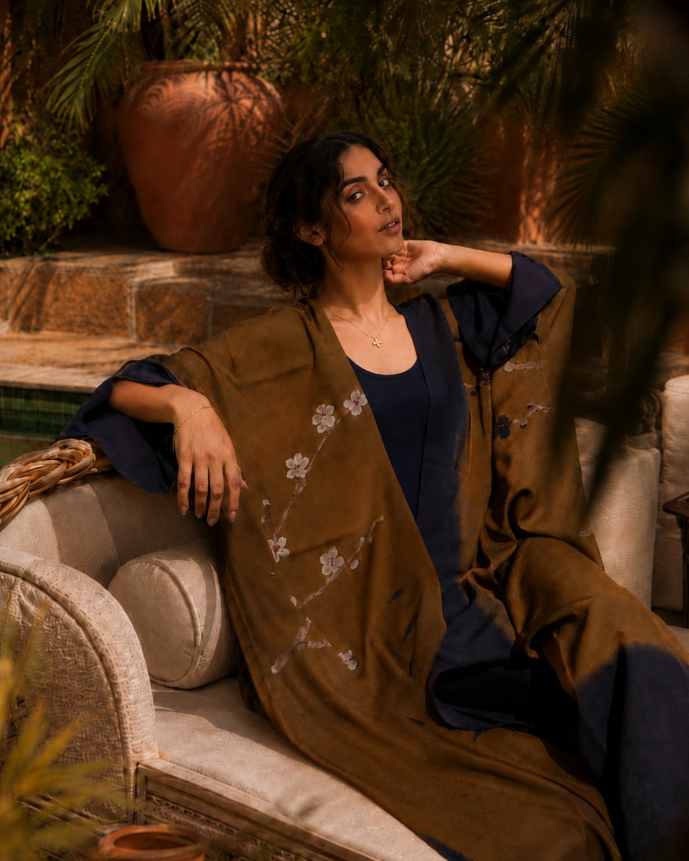 A woman sits on a cream-colored outdoor lounge chair in a garden setting, wearing a dark navy blue tunic and a draped brown shawl with delicate white floral embroidery.